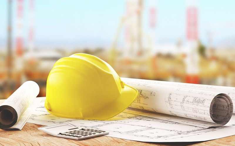 Un casque de chantier jaune et des plans de construction sont posés sur un bureau, avec un chantier en arrière-plan, symbolisant la planification et la gestion de projets avec un logiciel ERP.